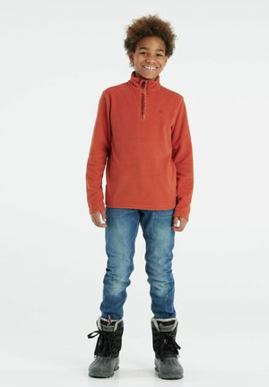 Fleece pullover in oranje met ritssluiting bij de kraag, gecombineerd met blauwe jeans en zwarte winterlaarzen met bontvoering en vetersluiting.