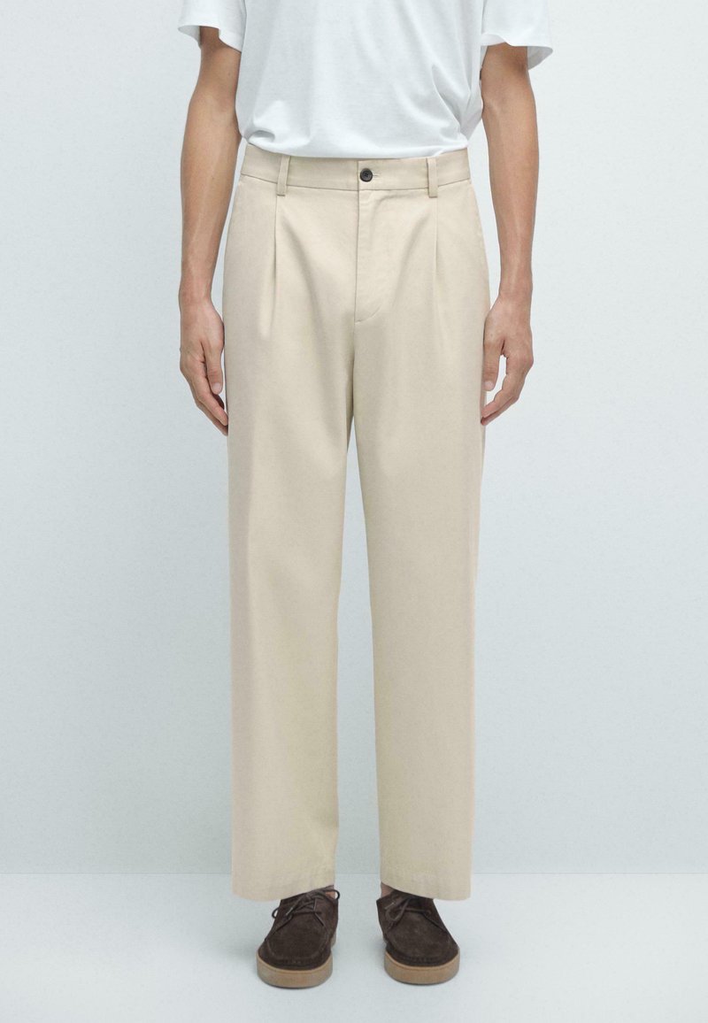 Beige pleated trousers made of soft fabric, featuring a button closure, side pockets, and a relaxed fit. Paired with dark brown shoes.