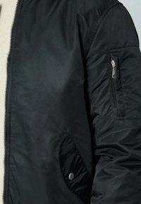 Veste bomber noire en matériau lisse et brillant. Dotée d'une fermeture éclair, de poches latérales et d'une poche zippée sur la manche. Texturée avec des poignets élastiques.