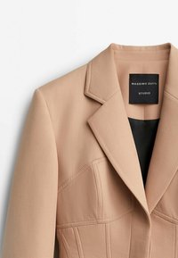 Blazer beige su misura con revers affilati, cuciture strutturate e foderato di nero. Presenta un'etichetta con la scritta "Massimo Dutti Studio."