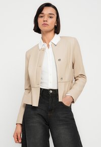 Μπεζ κοντό σακάκι με γιακά, κούμπωμα με ασημένια κουμπιά και λεία υφή, συνδυασμένο με λευκό πουκάμισο και σκούρο τζιν παντελόνι.