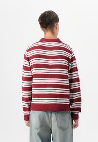 Jeune homme vu de dos portant un pull à manches longues rayé bordeaux et blanc et un jean denim bleu clair.