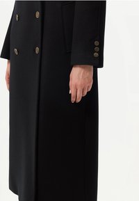 Svart dubbelknäppt kappa med guldfärgade dekorativa knappar på fram- och ärmslut, bärs av en person med synlig hand och handled.