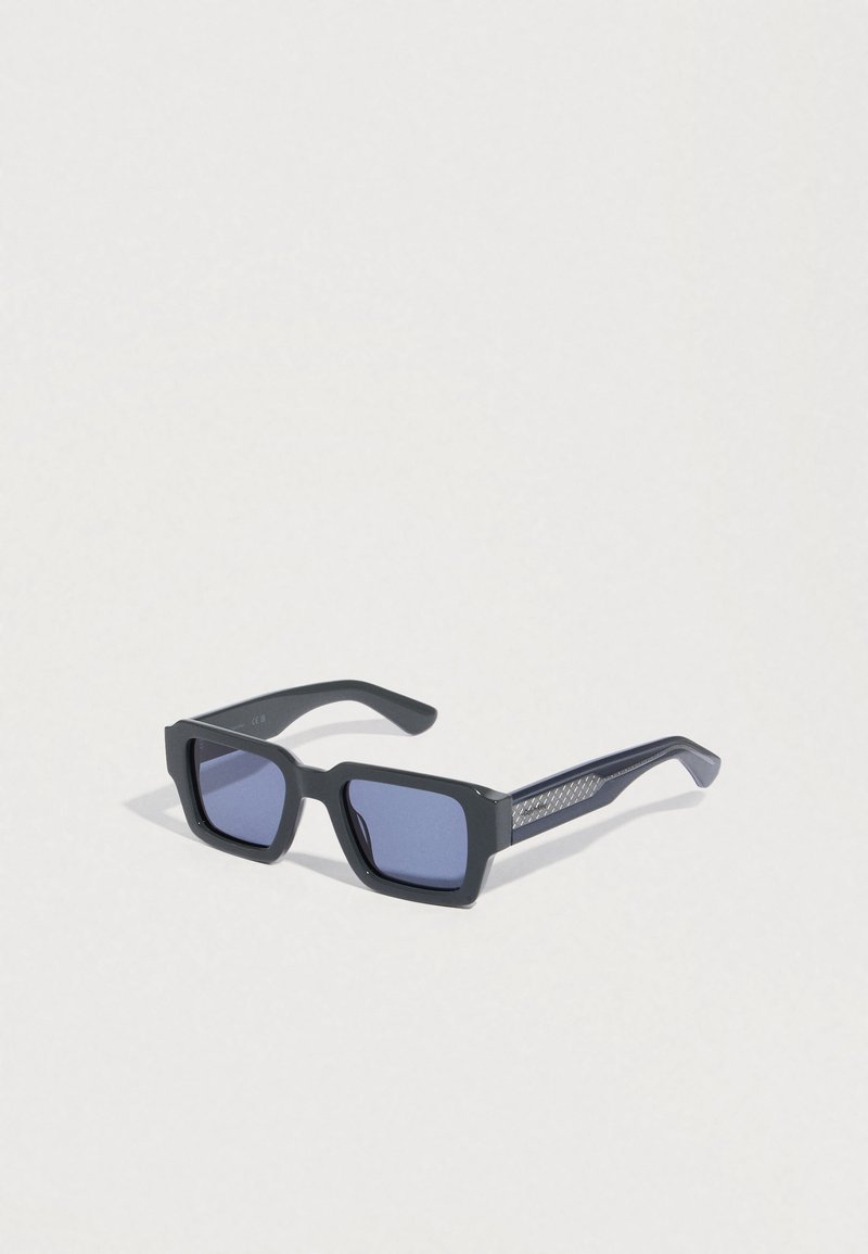 Zwarte rechthoekige zonnebril met donkere glazen. Voorzien van textuuraccenten aan de zijkanten, een dik montuur en gladde pootjes voor duurzaamheid.