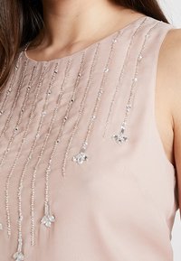 Růžový top bez rukávů se svislými řadami průhledných korálků a kovovými akcenty, vytvářející texturovaný, dekorativní efekt. Hladký povrch látky.