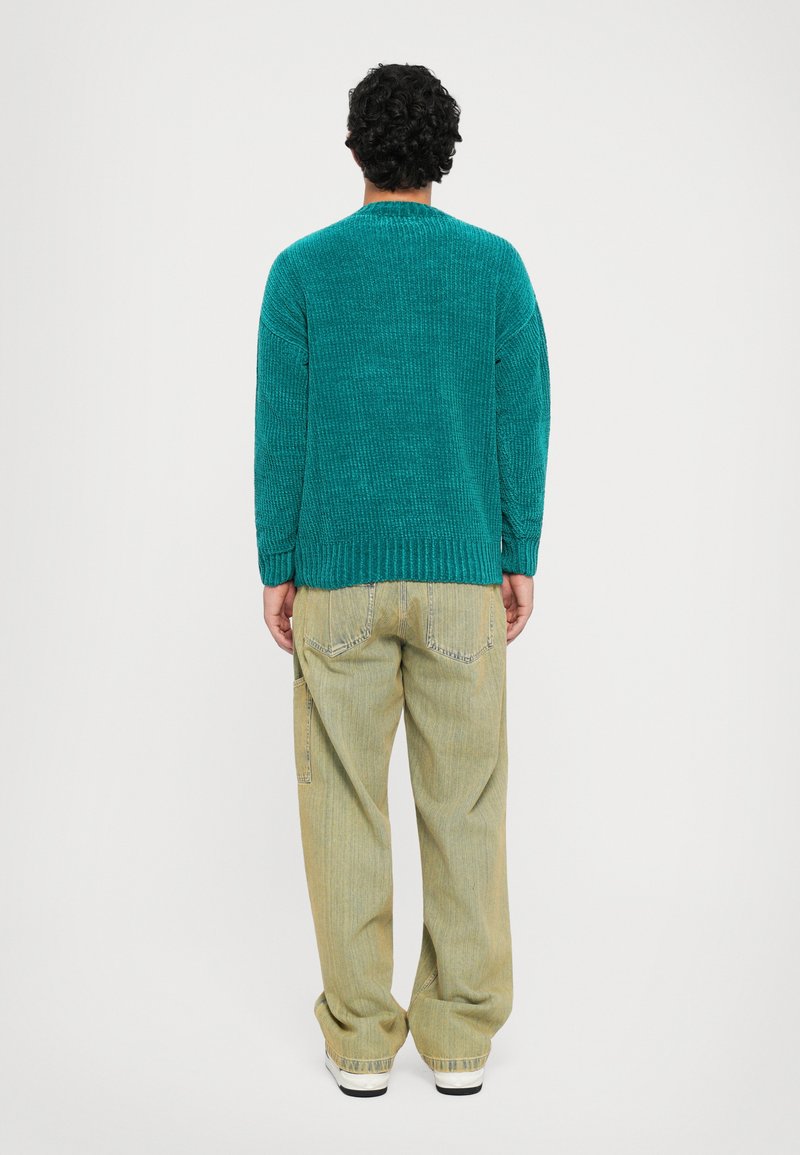 Man met krullend donker haar staat met zijn gezicht wegdraaien, gekleed in een teal gebreide trui, losse beige broek en witte sneakers, tegen een effen achtergrond.
