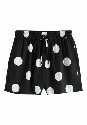 Manos levantando una camiseta negra, revelando unos pantalones cortos negros a juego con grandes lunares blancos, usados al aire libre bajo la cálida luz del sol.