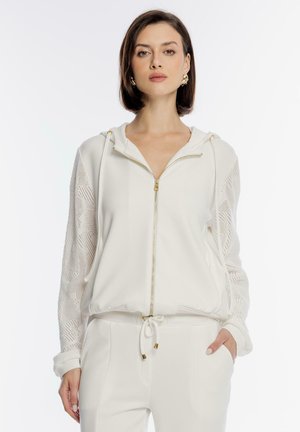 Kobieta ubrana w białą bluzę na zamek z koronkowymi rękawami oraz pasujące białe spodnie z troczkiem, stojąca na jednolitym białym tle.