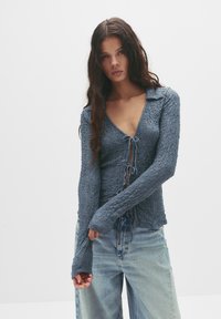 Blau strukturierter Langarm-Oberteil mit V-Ausschnitt, Kordelzug und schmalen Ärmeln. Kombiniert mit hellen Jeans.