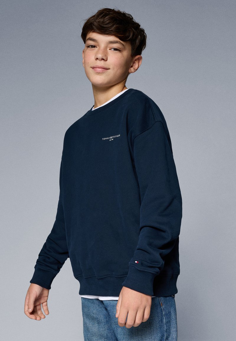 Felpa blu navy con collo a giro, polsini a coste e un piccolo logo sul petto. Indossata con jeans azzurri. Texture liscia.