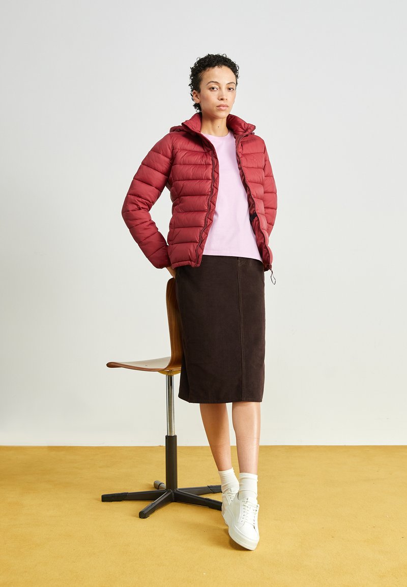 Chaqueta roja acolchada con capucha, camiseta rosa, falda lápiz marrón y zapatillas blancas, con un fondo amarillo y un telón de fondo neutro.