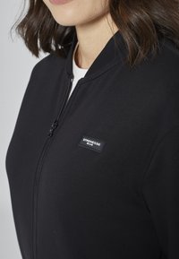 Veste noire zippée en tissu doux, avec un col côtelé, un patch logo sur la poitrine et une coupe ajustée.