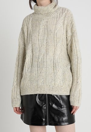 Femme portant un pull à col roulé beige en maille torsadée, associé à une mini-jupe en cuir noir brillant avec une fermeture éclair à l'avant.