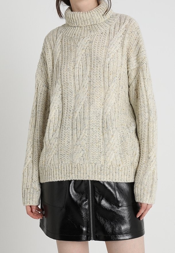 Femme portant un pull à col roulé beige en maille torsadée, associé à une mini-jupe en cuir noir brillant avec une fermeture éclair à l'avant.