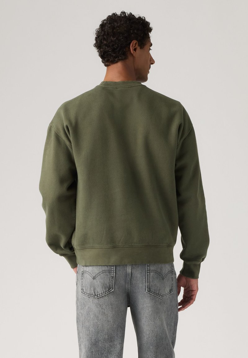 Uomo con capelli ricci che indossa una felpa verde oliva e jeans grigi chiari, visto da dietro su uno sfondo semplice.