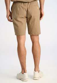 Pantalones cortos de tejido caqui con cinturilla elástica, que cuentan con un bolsillo trasero. Llevados con zapatillas deportivas de color claro.