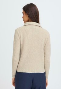 Beige ribbestrikket genser med brettet krage, lange ermer og en litt cropped hem, laget av et teksturert strikkemateriale.