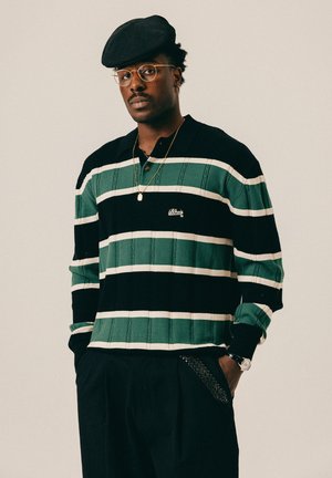 Mężczyzna w czarnym berecie, okrągłych okularach, zielono-czarnej pasiastym swetrze, czarnych spodniach z plecionym detalem, złotym naszyjniku i zegarku na nadgarstku, z rękami w kieszeniach.