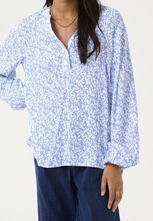 Femme portant un chemisier ample à manches longues bleu et blanc à motifs avec des boutons, associé à un pantalon bleu foncé et un collier chaîne en or.