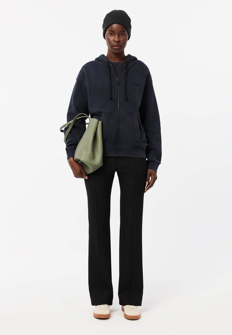 Marinefarbener Zip-Hoodie, schwarze Tailored-Hosen und weiße Sneakers. Trägt eine grüne Lederhandtasche und eine dunkle Strickmütze.