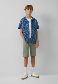 Camisa azul de manga corta con patrón de hojas, camiseta blanca debajo, pantalones cortos verdes, calcetines blancos y zapatos blancos con suelas beige.