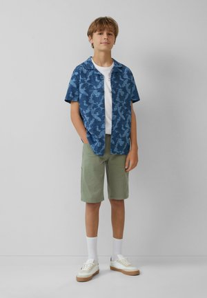 Korte mouwen blauwe shirt met bladpatroon, witte t-shirt eronder, groene shorts, witte sokken en witte schoenen met beige zolen.
