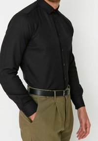 Zwarte lange mouwen shirt met een spits kraag, knoopsluiting aan de voorkant en een aansluitend ontwerp, gecombineerd met olijfgroene broek en een zwarte riem.