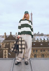 Grön och vit randig polo skjorta, gråa sweatpants, svarta loafers med blommönster, hållande en rutigt jacka, stående på ett tak.