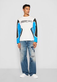 Long-sleeve white t-shirt with black and blue accents, featuring the text "981sports REPLAY." Worn with distressed blue jeans and white sneakers.