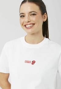 T-shirt en coton blanc avec un logo graphique rouge des Rolling Stones, comprenant leur design emblématique de lèvres et langue, manches courtes.