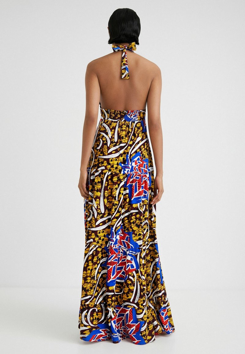 Femme aux cheveux courts et foncés portant une robe longue dos nu à bretelles nouées avec un motif abstrait et coloré en jaune, rouge, bleu, blanc et marron.