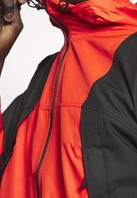 Červená a černá mikina s kapucí se zapínáním na zip, nastavitelným stahovacím šňůrkem a měkkou texturou s kontrastními panely.