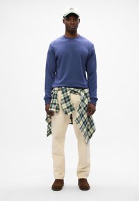 Vijolična majica s kapuco, kremne hlače, zeleni črtasti flanel vezan okoli pasu, rjavi čevlji in svetlo zeleni kapica s podrobnostmi logotipa.