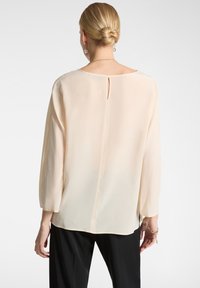 Blusa beige chiaro con maniche lunghe leggermente a sbuffo, apertura a chiave sulla schiena e un tessuto morbido e leggero, abbinata a pantaloni neri.