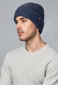 Gebreide muts in donkerblauw, met ribbeltextuur. Aangehechte label aan de zijkant. Geverfd met een lichtgrijze ribbeltrui.