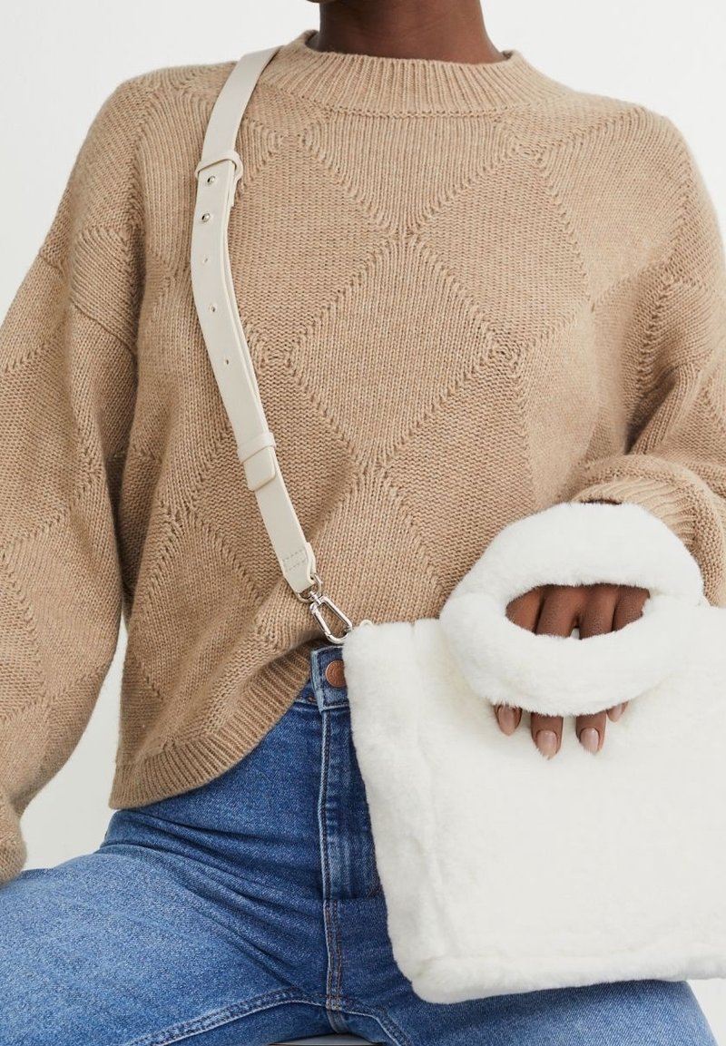 Personne portant un pull en maille beige texturé et un jean bleu, tenant un sac à main blanc duveteux avec une poignée ronde et une sangle ajustable.