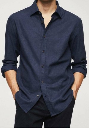 Uomo che indossa una camicia a maniche lunghe di colore blu scuro con le maniche arrotolate, mani casualmente nelle tasche, su uno sfondo semplice.