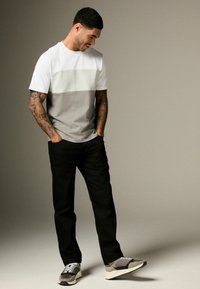 T-shirt à manches courtes, bicolore en blanc et gris, associé à un pantalon noir. Le mannequin porte des baskets grises et blanches.