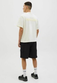 Tricou din bumbac de culoare crem deschis, cu inscripția "MALIBU" imprimată în galben pe spate, asortat cu pantaloni scurți negri din material cargo și adidași negri.