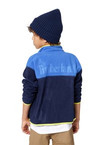 Blå og marineblå fleecejakke med "Timberland"-logo, ribbestrikkede mansjetter og en gul aksent ved kanten. Kombinert med en marineblå lue.