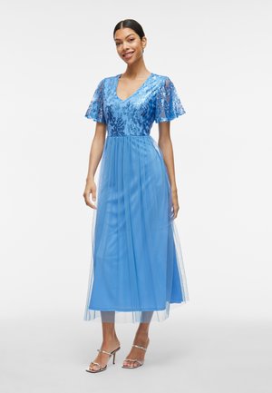 Robe bleue avec un corsage pailleté, des manches transparentes et une jupe fluide en tissu léger. Accessoirisée avec des talons argentés à lanières.