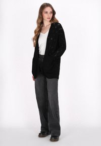 Cárdigan negro de punto con apertura frontal, que presenta un patrón texturizado. Combinado con pantalones de mezclilla negros de cintura alta y zapatos oscuros.