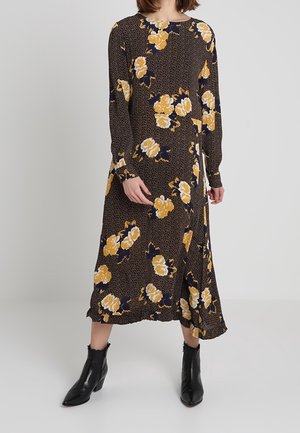 Woman wearing a long-sleeve, ankle-length black dress with yellow floral print and black ankle boots, standing against a plain background.