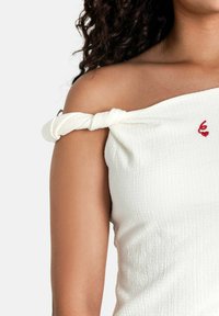 Witte off-the-shoulder top met knoopdetail op de band, textuurstof en een kleine rode geborduurde accent op de borst.