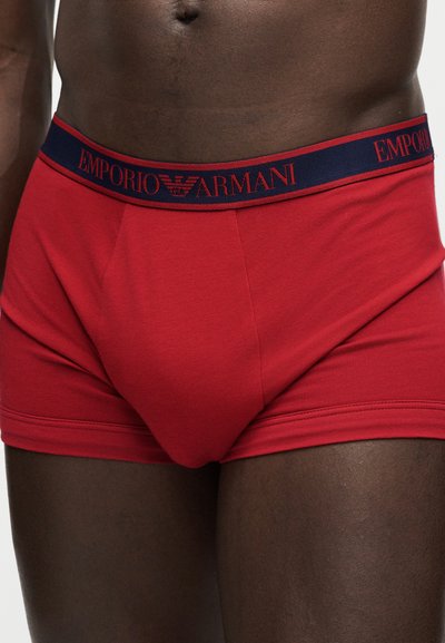 Közeli kép egy személyről, aki piros Emporio Armani boxeralsót visel tengerészkék derékpánttal és piros logós felirattal.