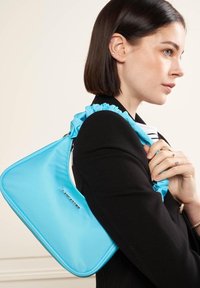 Sac à main synthétique bleu clair avec une forme triangulaire unique et une anse froncée. Présente un logo argenté "LANCASTER".