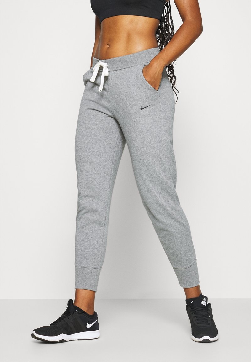 Femme portant un pantalon de survêtement Nike gris avec un cordon blanc, un crop top noir et des baskets Nike noires, debout devant un fond uni.