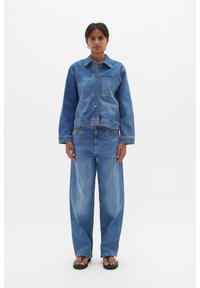 Modra denim jakna z sprednjim gumbnim zapiranjem in žepek, v kombinaciji z širokimi modrimi denim hlačami. Model stoji ob beli ozadju.