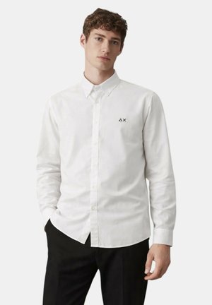 Giovane uomo che indossa una camicia bianca a maniche lunghe con un piccolo logo nero sul petto e pantaloni neri, in piedi con una mano in tasca.