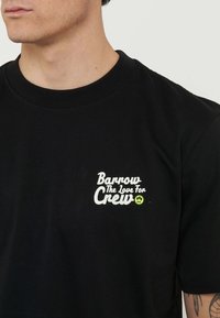 Hombre con camiseta negra con texto blanco "Barrow The Love For Crew" y pequeña cara sonriente verde en el pecho izquierdo.